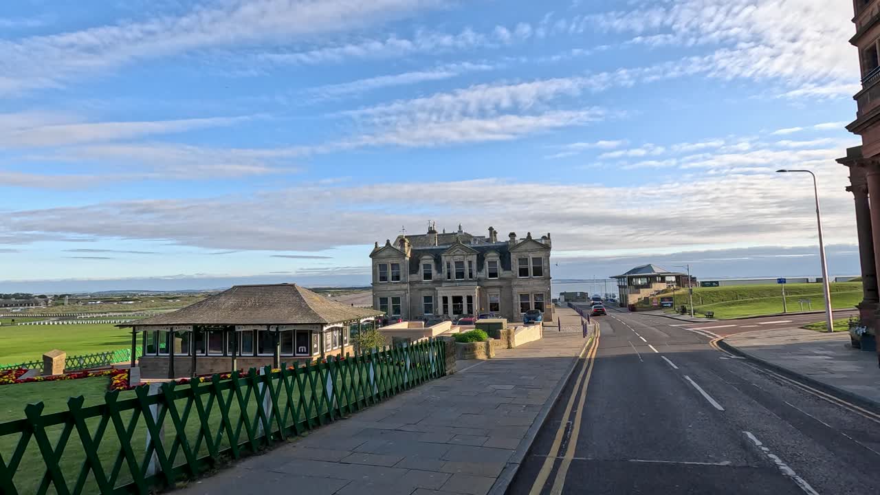 Person walks along road toward iconic golf clubhouse, under bright daylight, with scenic seaside views