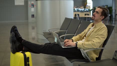 Un-Joven-Aburrido-Está-Sentado-En-El-Aeropuerto,-Esperando-Que-Salga-El-Avión-Y-Usando-Una-Computadora-Portátil