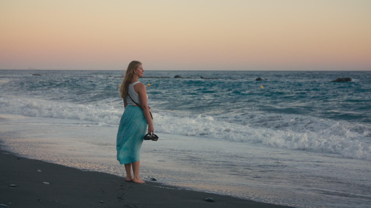 A barefoot woman in a blue skirt and white top stands calmly by the sea, holding her shoes, gazing out at the horizon as the breeze lifts her skirt. Wide shot