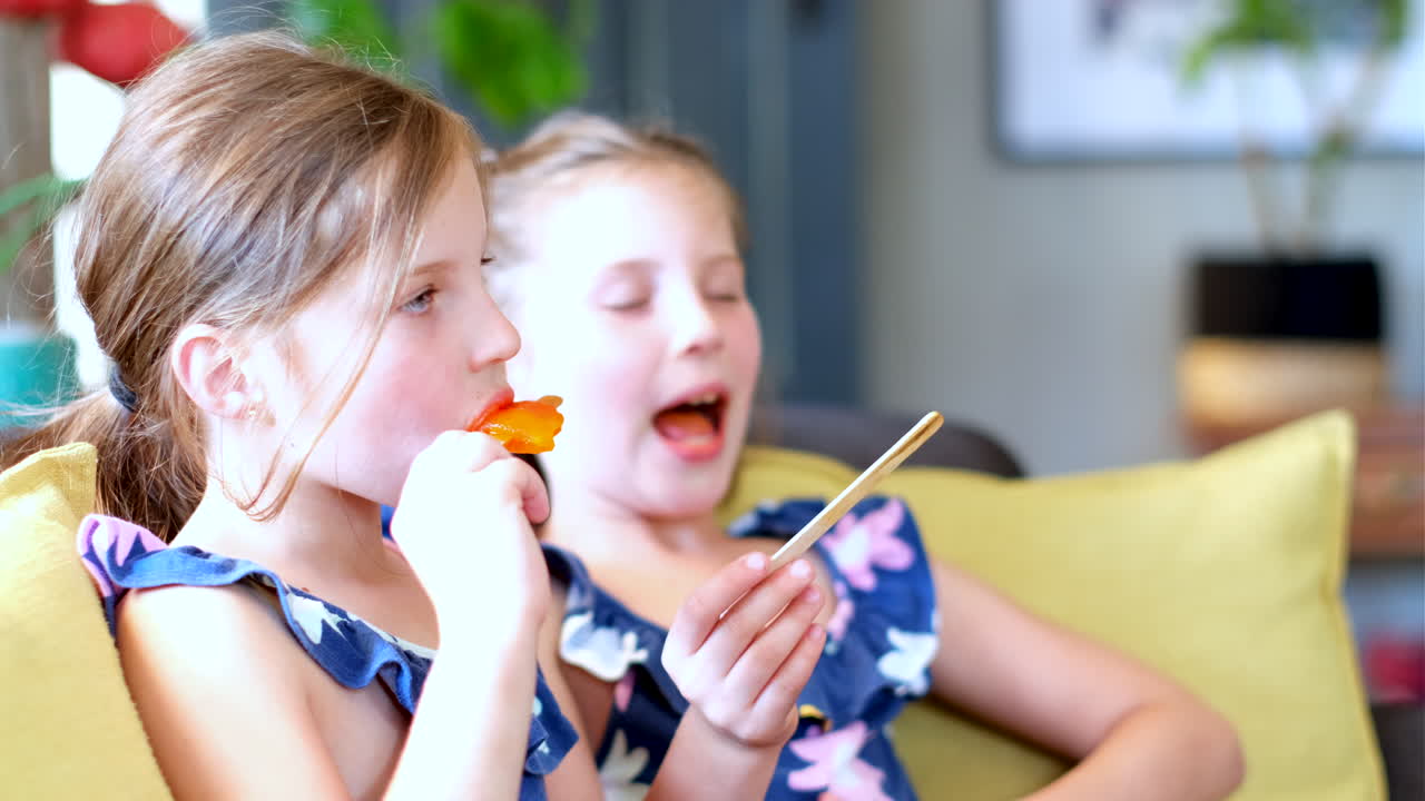 Caucasian fraternal twin girls enjoy orange ice pops, close-up side lighting