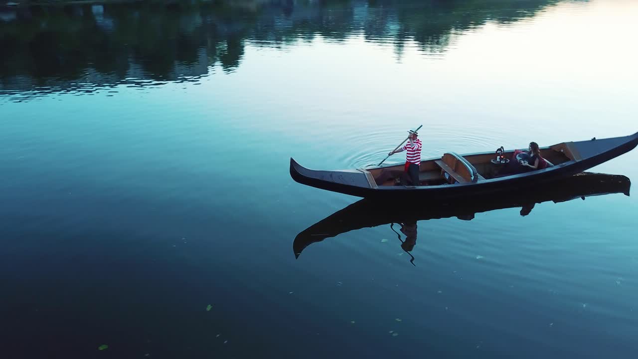 Aerial view of a boat floating in the river at sunset. Young woman incarnival mask sitting in gondola and a gondolier standing with an oar. Camera moves around.