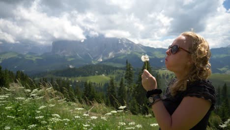 super slow mo of a blonde girl blowing a dandelion flower in the wind in a beautiful nature setting outdoor in the summer, val gardena, alpe si siussi