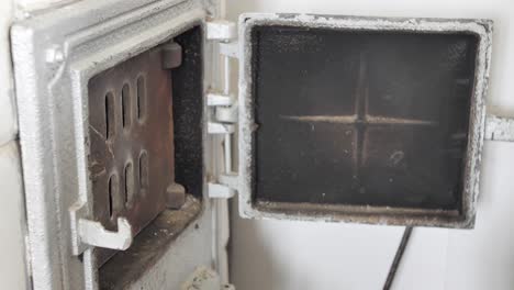 man opens the door of old home tiled stove and puts a firewood inside