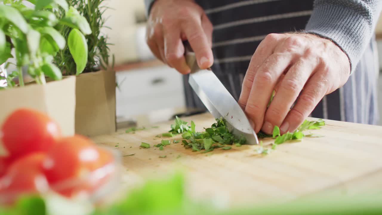 sección media de un hombre caucásico de pie en la cocina, cocinando la cena, cortando albahaca