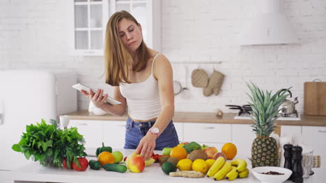beautiful woman looking into tablet on the light kitchen background. young housewife chooses necessary organic fruit and vegetables for recipe to lose weight. vertical video