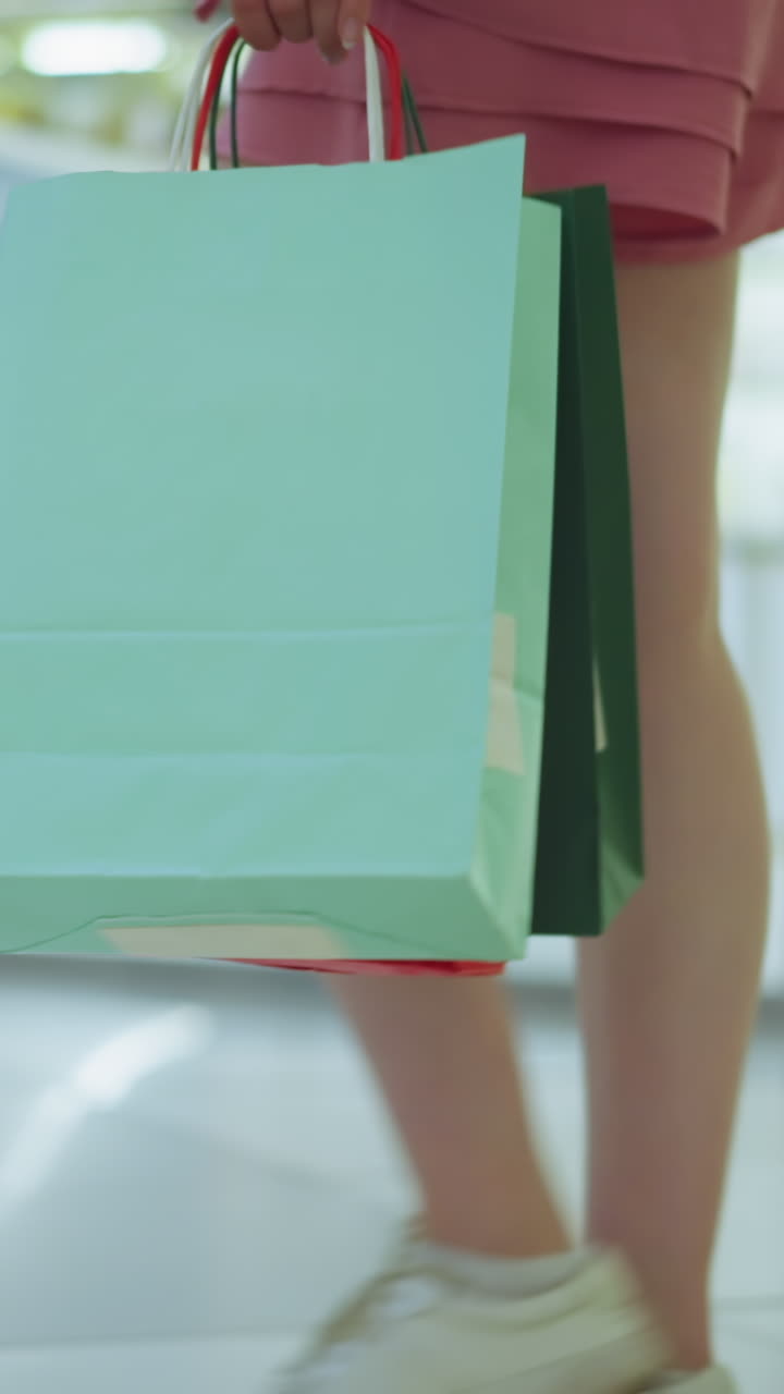 밝게 조명된 쇼핑몰을 걸어가면서 다채로운 쇼핑 가방을 들고 있는 여자의 다리 시각, 그녀가 바닥을 가로질러 웅장하게 움직일 때 배경에 보이는 상점 품목의 약간의 흐릿함