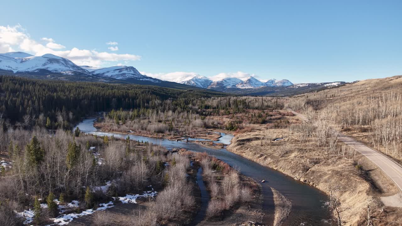 Riprese con drone seguono un fiume serpeggiante attraverso un'ampia valle brulla, costellata di alberi e arbusti sempreverdi, con montagne innevate all'orizzonte vicino al Glacier National Park in Montana.