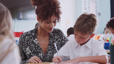 Woman-Elementary-School-Teacher-Giving-Male-Pupil-Wearing-Uniform-One-To-One-Support-In-Classroom