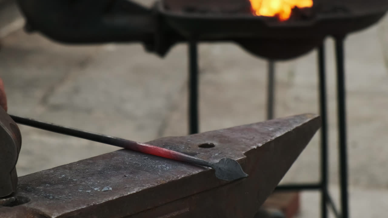 High-angle static close-up of a blacksmith hammering a red-hot spearhead on an anvil; glowing steel and precise strikes in a gritty workshop backdrop