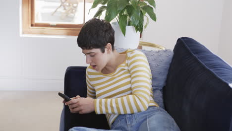 happy caucasian woman lying on sofa, using smartphone and laughing in sunny house