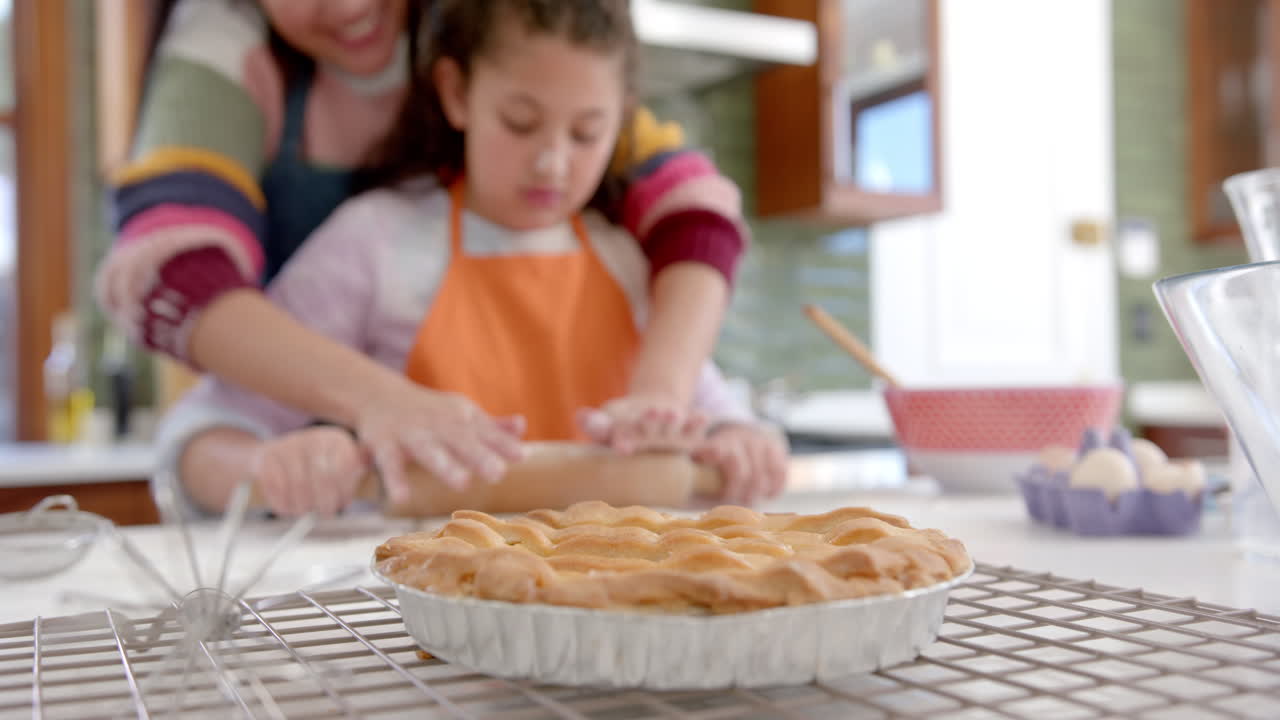 madre y hija bi-raciales felices rodando la masa y sonriendo en la cocina soleada