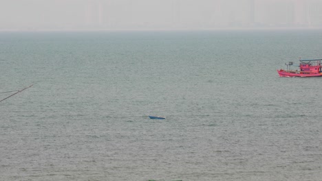 Ein-Kleines-Blaues-Boot,-Das-Mit-Einem-Vorbeifahrenden-Thailändischen-Fischereifahrzeug-Im-Hintergrund-Vor-Der-Küste-Von-Pattaya,-Thailand,-Verankert-Ist