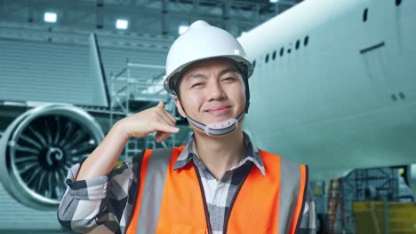 close up of asian male engineer with safety helmet standing with aircraft in the hangar. smiling to camera and making call me gesture while aircraft maintenance