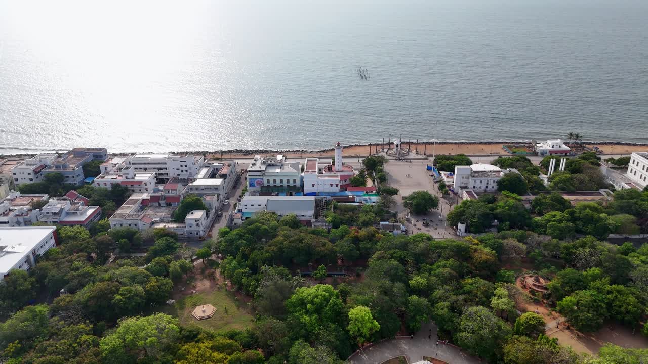Filmato aereo di un lussureggiante parco verde che si trasforma in un'area urbana costiera. In primo piano, è visibile un parco tranquillo con sentieri tortuosi e un gazebo bianco centrale. Statua di Gandhi a Pondicherry.