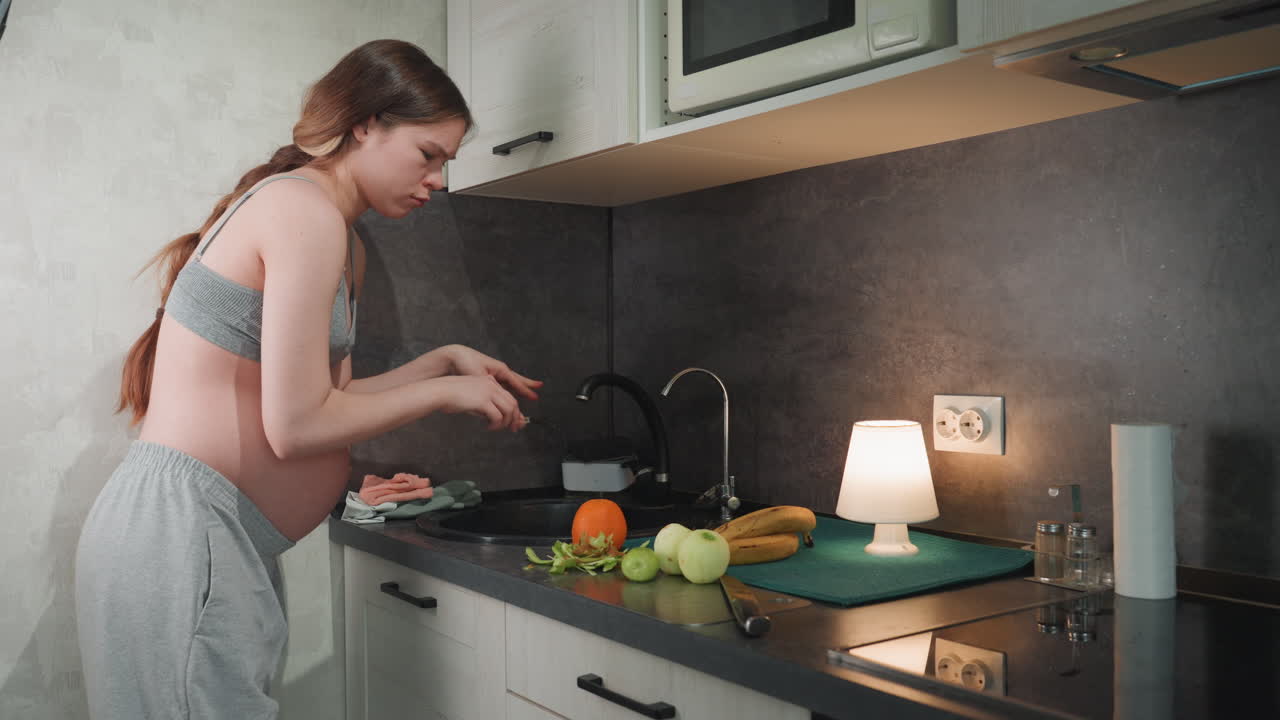 Donna caucasica incinta che taglia verdure, luce soffusa della lampada, piano di lavoro con mele, arance, carote e tagliere, abbigliamento casual da casa, movimenti delicati, che condisce e sistema l'insalata, prenatale.