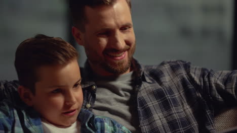 Smiling-father-and-son-sitting-on-couch-with-computer.-Dad-stroking-kid-boy-head