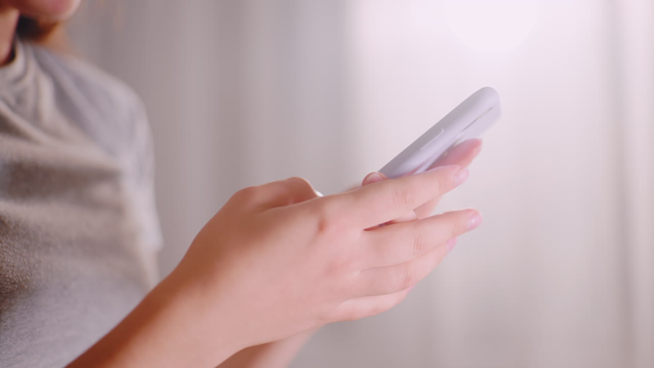 Close up of lady holding smartphone with both hands, chatting and texting as soft sunlight shines through background curtain, focus on natural gesture and casual lifestyle