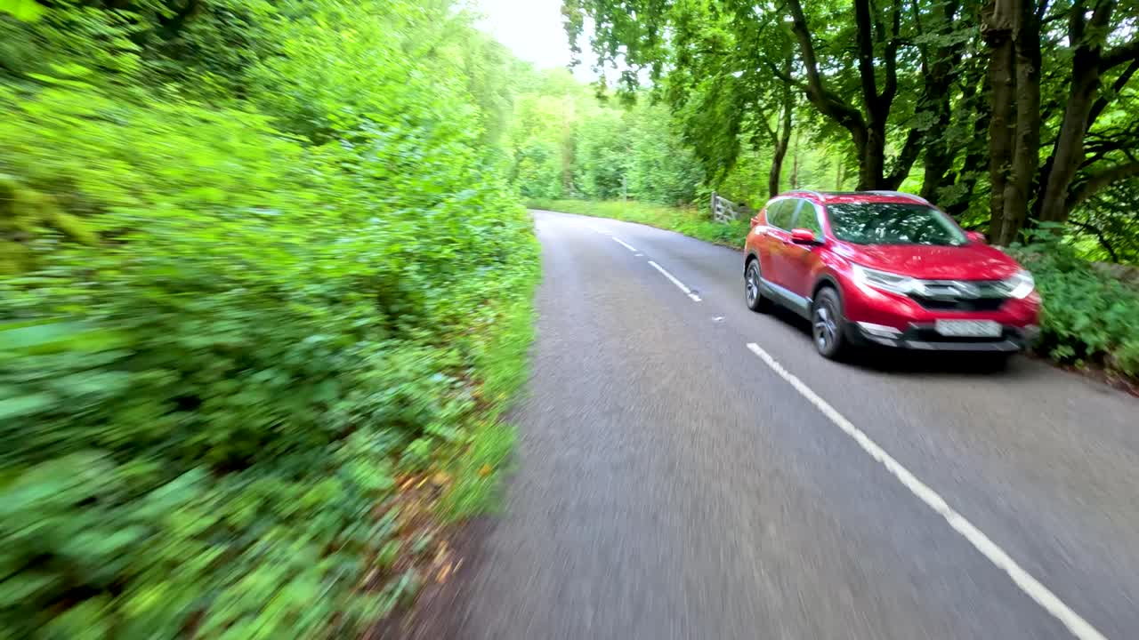 A vehicle travels along a winding, tree-lined country road in bright daylight, passing other cars and dense green foliage, captured with smooth forward camera movement