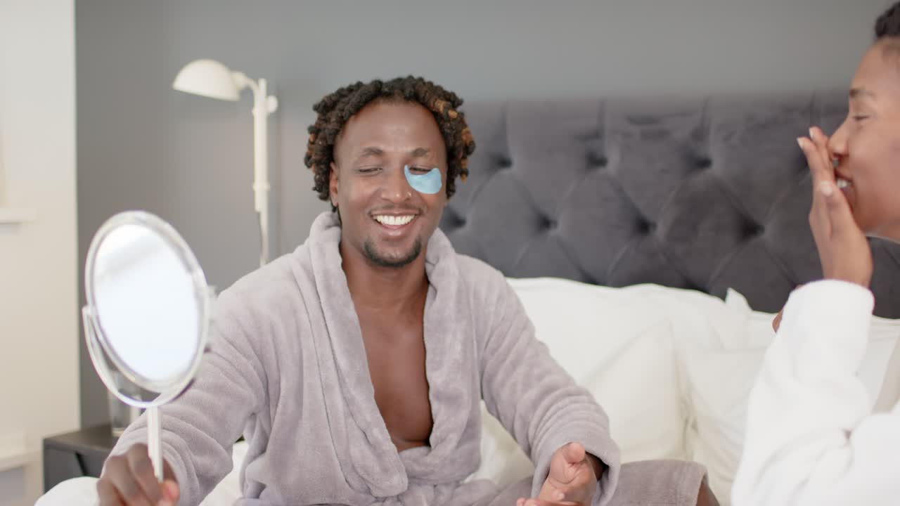 African American couple sitting on bed holding mirror after seeing under-eye gel patch, laughing