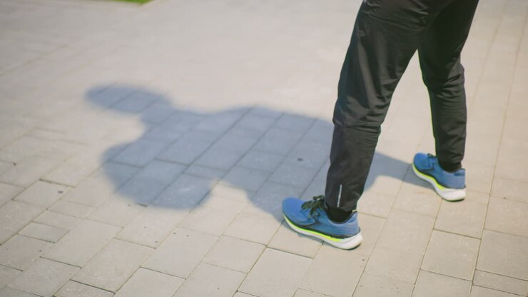 pavement shadow of pedestrian standing still, closeup of legs and sneakers, long soft silhouette stretching across tiled plaza, morning sunlight casting clear outline, minimal color palette, calm