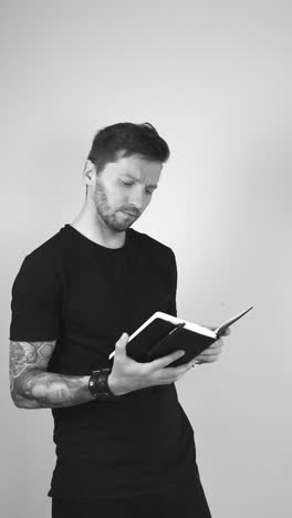 grayscale footage of a tattooed man opening up a journal diary or a little black notebook and starts reading it while flipping the page at some point. man is with a thinking face and investigating.