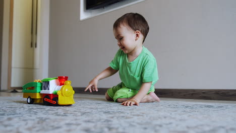 smiling caucasian boy sits on the floor playing with toy. happy healthy child pushes his train.