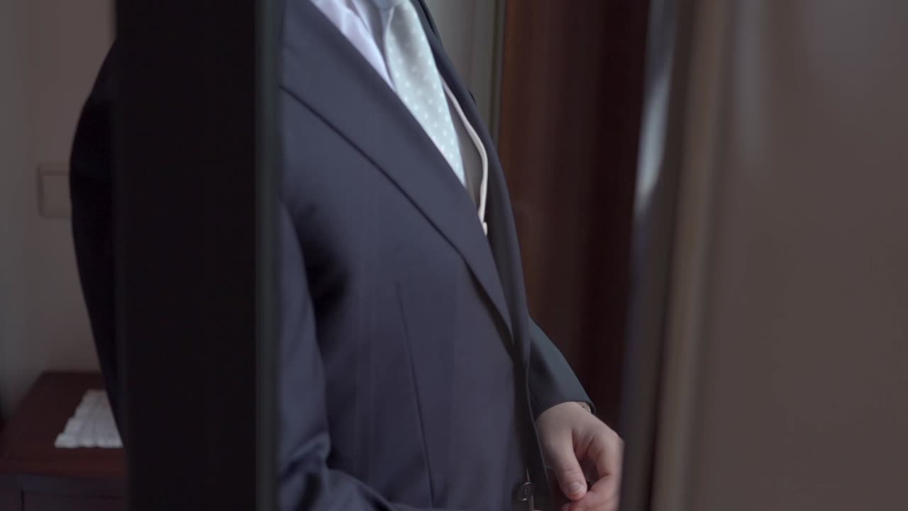 Man buttoning dark suit jacket in front of a mirror with a light green tie