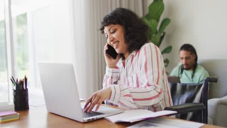 feliz mujer biracial en silla de ruedas hablando por teléfono usando computadora portátil, con compañero masculino en la sala de estar