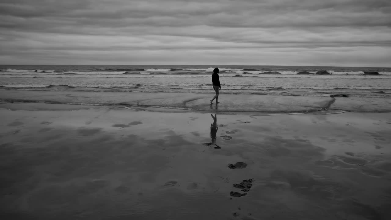Black and white video still of a lone figure walking on a beach