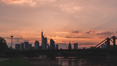 silhouette of frankfurt skyline against a sunset sky with clouds and bridge over river, timelapse