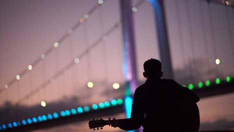 a young man playing the guitar to himself whilst sat on a small beach overlooking a pretty and colourful bridge in a city in america