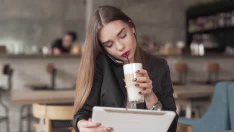 busy young professional woman talks on her cellphone, uses her tablet, and drinks coffee while sitting in a café