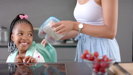 biracial mom helps daughter with backpack in kitchen for school.