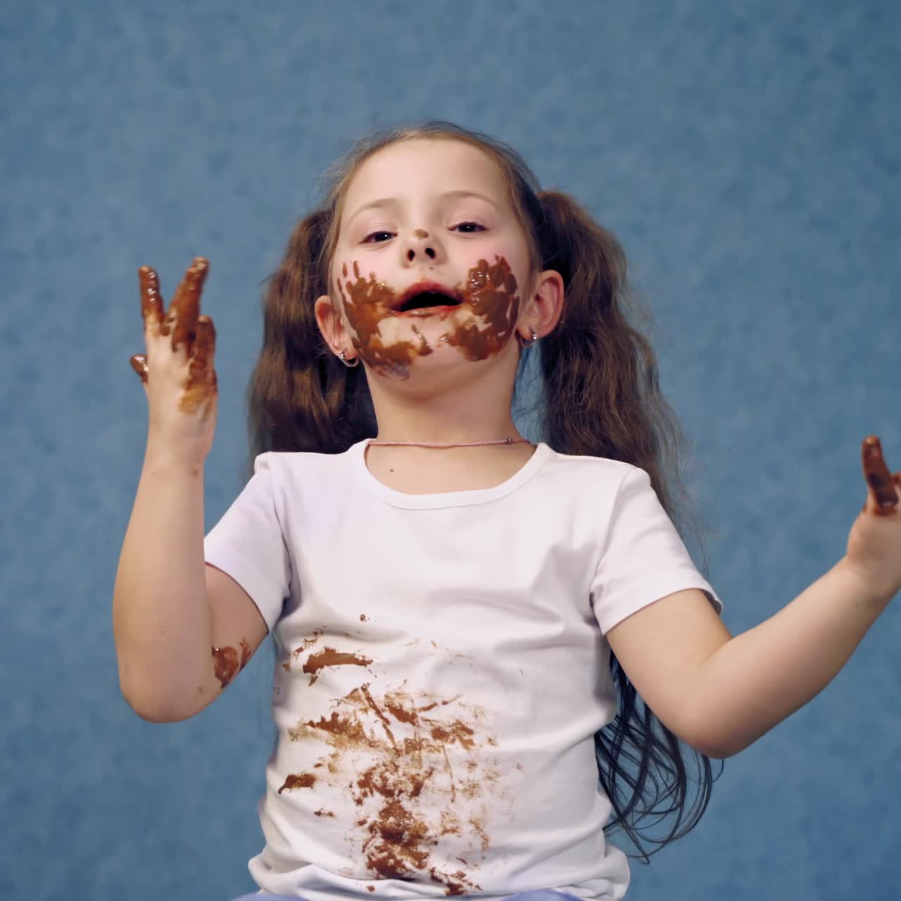 Funny little girl in smeared chocolate on face and hands. Cute joyful child licking her fingers with tasty leavings of chocolate. Dirty girl on blue background.