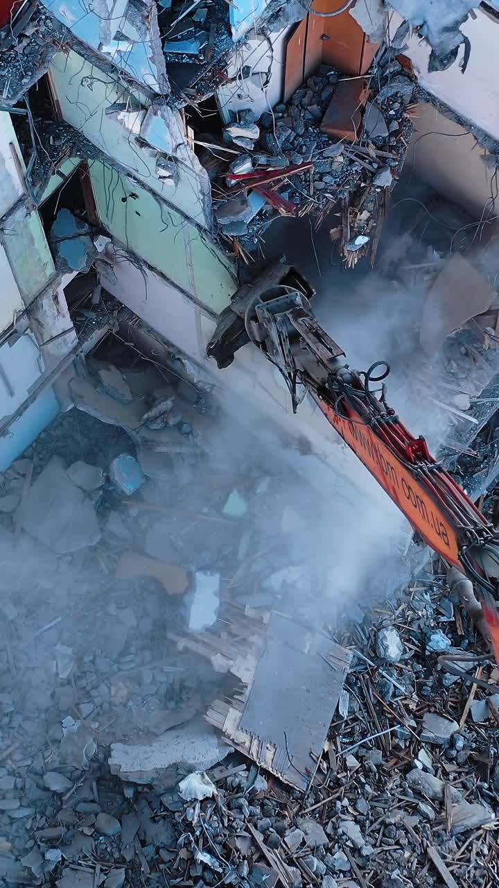 Huge building being demolished with the help of a tractor. Excavator with a long claw breaking walls of the ruined building. View from the above. Vertical video