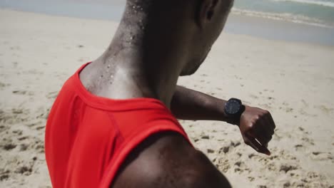 African-american-man-checking-his-smartwatch,-taking-break-in-exercise-outdoors-by-the-sea