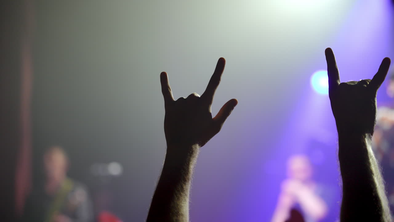 Energetic music concert crowd experiencing collective excitement, raising hands and making enthusiastic gestures under vibrant purple stage lighting with blue balloon floating overhead