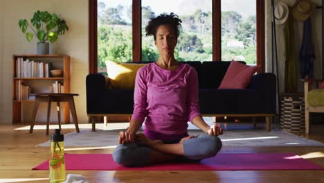 Mixed-race-woman-practicing-yoga-and-meditating-while-sitting-on-yoga-mat-at-home