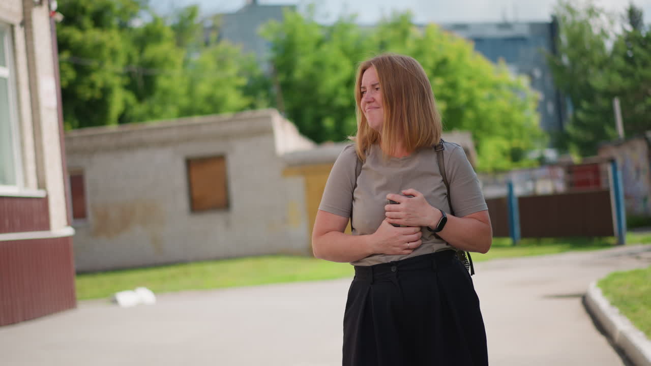 Woman with golden hair standing on sidewalk using smartphone on sunny day, casual clothing, green trees and graffiti wall in background, calm facial expression capturing everyday