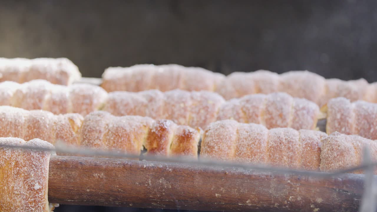 Rows of sugar-coated chimney cakes rotate on a wooden spit over an outdoor grill, with a hand occasionally adjusting them. Natural daylight, close-up perspective