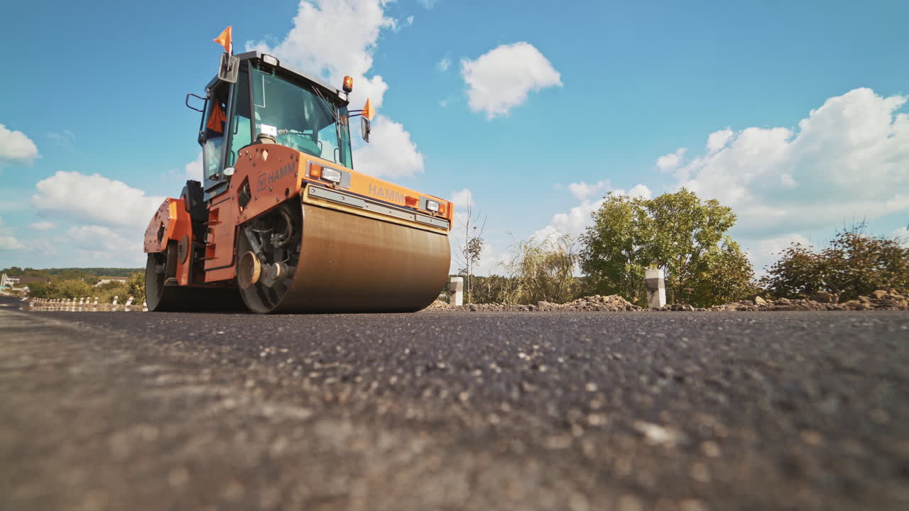 Orange roller machine pressing hot asphalt on the road in summer. Heavy compactor machine making new asphalt on the background of blue sky and green trees. Road resurfacing.