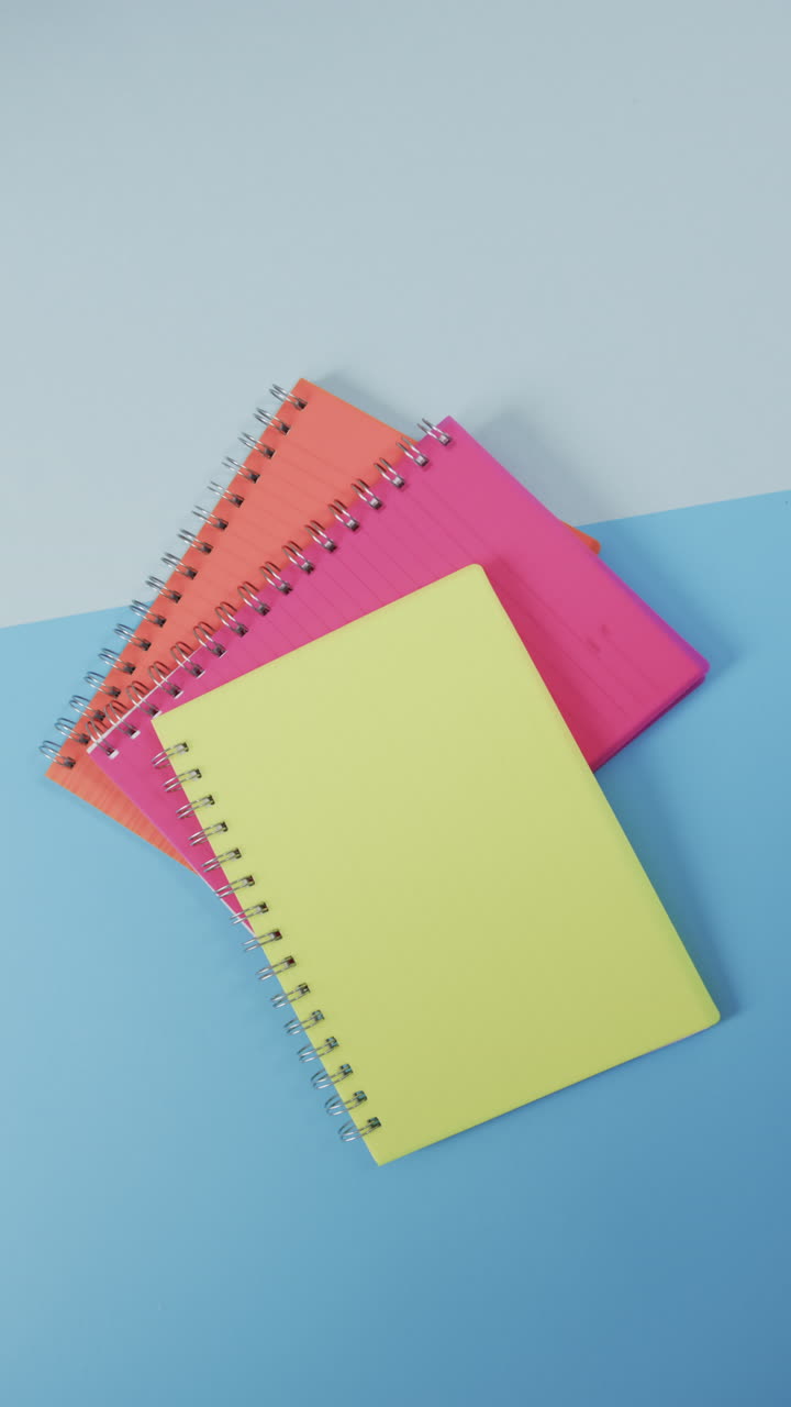 Vertical video of overhead view of colourful notebooks on blue background, in slow motion