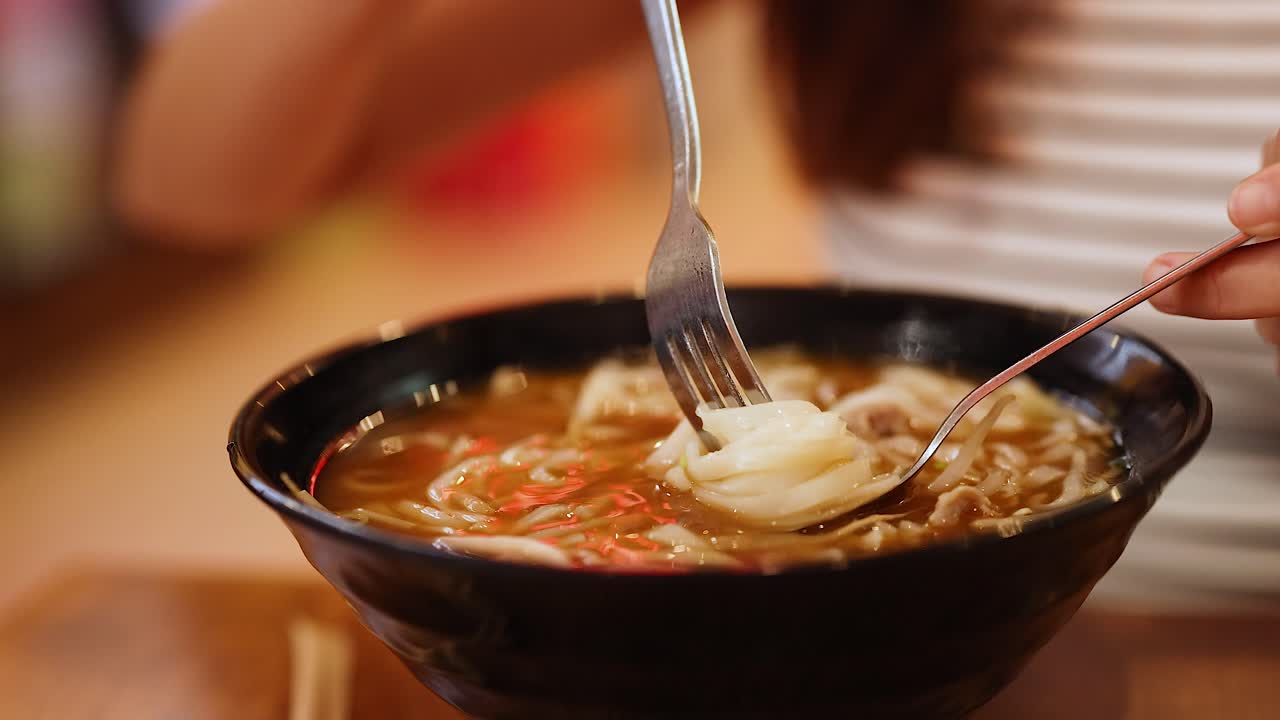 A person savors a bowl of Vietnamese pho, using a fork and spoon. Warm lighting enhances the cozy dining atmosphere