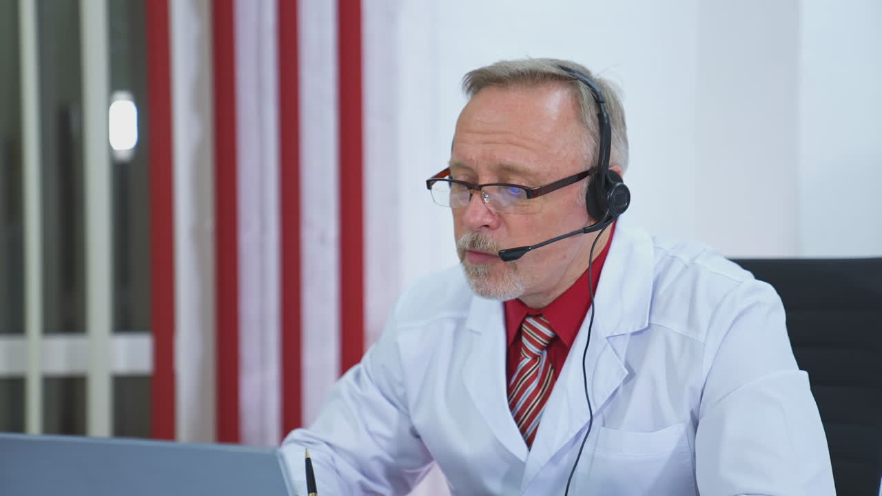 Remote consultation. Senior doctor in medical coat talking online and writing in medical office. Medical specialist consults patient remotely using laptop and headset.