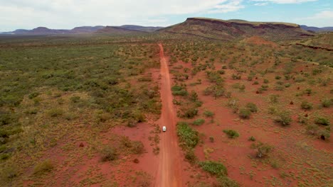 aerial view of a vehicle traveling on a long dirt road through a vast arid outback landscape with distant mountains.