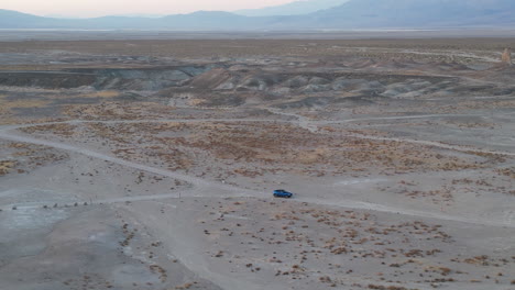 an aerial view of the side of a blue car as it turns at an intersection of dirt roads in the desert landscape at sunset