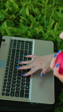 dama rusa acostada en la hierba con gafas en la cabeza escribiendo en la computadora portátil con la mano derecha mientras sostiene una manzana parcialmente comida en la mano izquierda, un entorno al aire libre relajado rodeado de vegetación vibrante