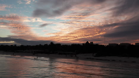 As-the-sunsets-in-Tulum,-Mexico,-vacationers-walk-along-the-beach-silhouetted-in-the-foreground