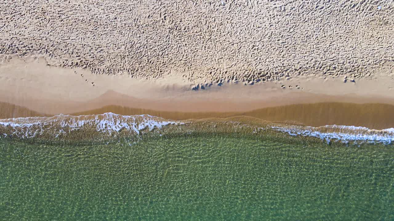 The sand looks brown and has a few visible footprints. The water is blue green, with waves crashing on the shore.