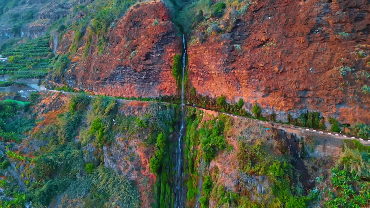 Jaw-dropping view of a high mountain with a road in the middle. Thin waterfall flows by the red rocks. Aerial perspective on the nature of the Madeira Islands, Portugal. Drone footage.